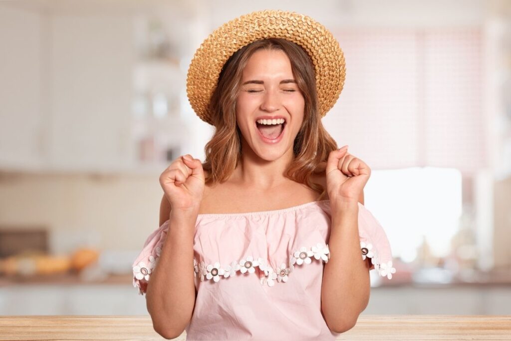 A joyful woman in a straw hat and a pink off-the-shoulder top celebrating with a wide smile and closed eyes, symbolizing a successful or easy day thanks to 'mom hacks.'