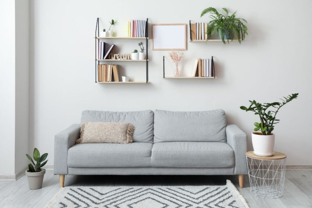 A small, stylish grey sofa with wall-mounted shelves above it, showcasing books and plants. This affordable vertical storage hack is a great space-saving idea for on-the-go moms to maximize space and create a functional, calm home